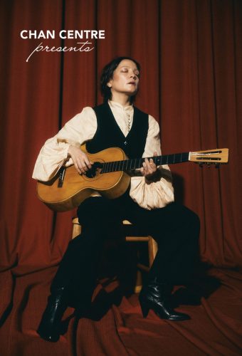 Natalia Lafourcade holding a guitar against a dark red backdrop, eyes closed, seated in a chair